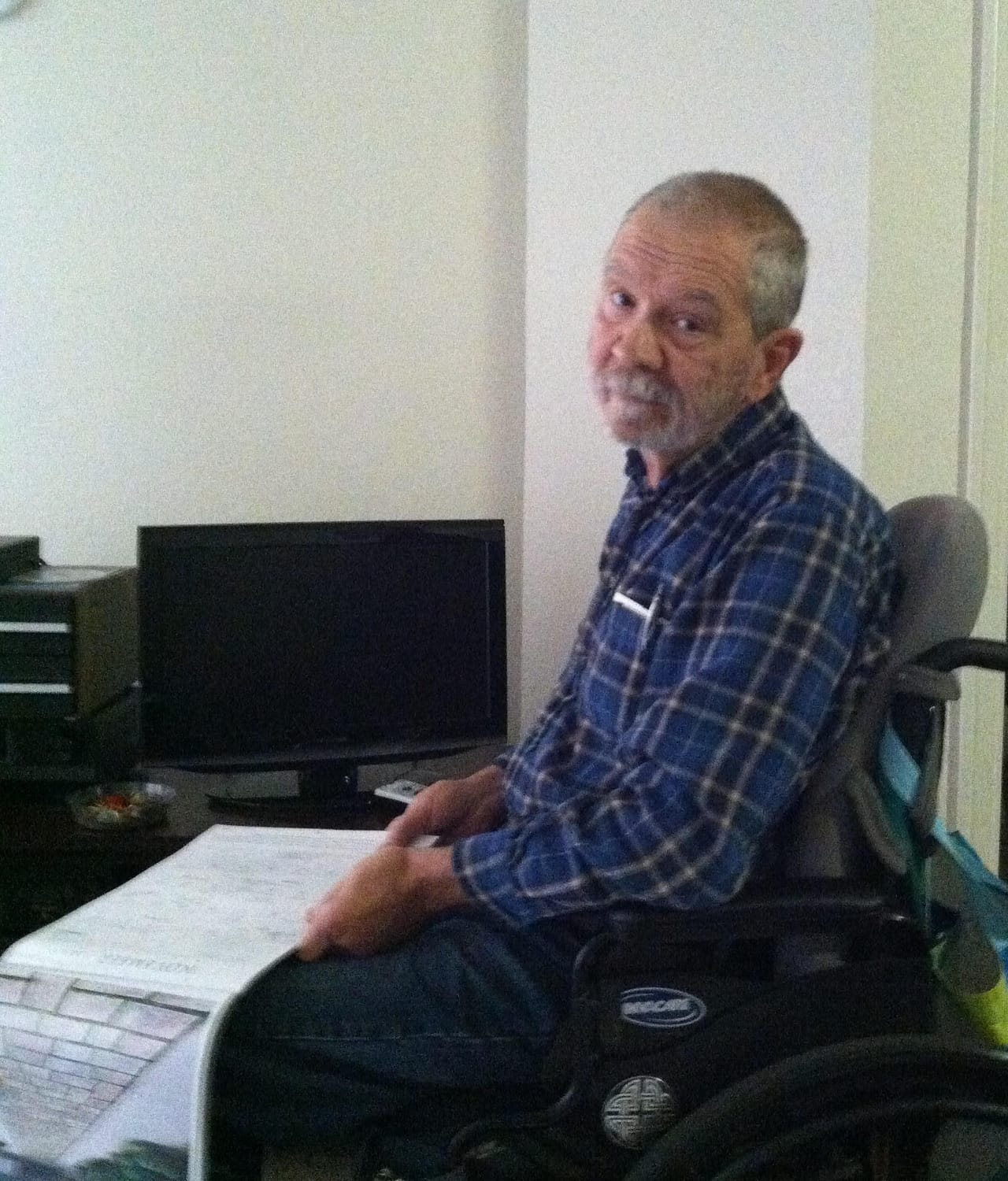Ed Milewski, champion of Independent Living, is pictured seated in a wheelchair with his body turned to the side, but his face looking at the camera. He is wearing a blue plaid shirt and black pants. The background is simple white walls, with a computer monitor directly behind him. In his hands he is holding a folded pad of paper.