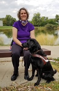 Beth Gustin is sitting on a bench outside. She is wearing a flowered scarf around her neck, a purple shirt, and black pants. She is smiling, and has chin-length curly hair. In front of her is her black lab Manolo, who is her guide dog. Manolo is wearing a brown leather harness.