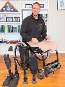 Craig Towler, Community Advocate for CPWD. Image description: Craig sits in his wheelchair. He has short brown hair, and is wearing a black shirt and tan pants. On the ground in front of him are two pairs of prosthetic legs. One set has sneakers on the end, the other set ends in flat grey pads. Behind him is a bookshelf with various photos and books.