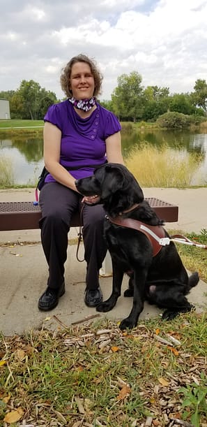 Beth Gustin and her service dog Manolo. Image description: Beth Gustin is sitting outside on a flat bench. She is wearing a purple t-shirt, floral scarf around her neck, and black pants. Her curly brown hair is chin-length. In front of her is sitting her black lab Manolo. He is wearing a harness with a protruding handle.