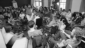 Demonstrators sit in at the offices of the Health, Education and Welfare department in San Francisco on April 9, 1977, advocating for civil rights rules for the disabled to be signed. Image description: In this black and white image, we see dozens of people sitting in a circle, about half of them in wheelchairs. In the center of the circle stands a woman talking into a microphone.
