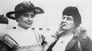 Black and white photo of Helen Kaller and Anne Sullivan. Keller stands on the left and is wearing a brimmed hat with a flower and a white collared blouse. Her hand is resting on Anne Sullivan's mouth, who stands to her right, looking at Helen. She is wearing a dark pill box hat, a white blouse, and dark jacket.