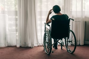 Image description: A woman in a wheelchair sits facing a window with closed drapes. Her back is to the camera. Her hand is to her head as if she is distressed.
