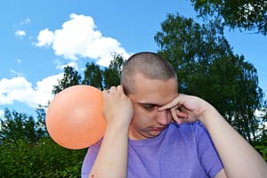 The image is of an autistic boy who appears agitated. He is outside with blue sky behind him. He is holding a balloon close to his ear, while his other hand is pressed to the side of his face with one finger over his eyes. He is wearing a purple shirt and had a shaved head.