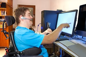 Image description: A young man with infantile cerebral palsy caused by a complicated birth sitting in a multifunctional wheelchair, using a computer with a wireless headset, reaching out to touch the touch screen.