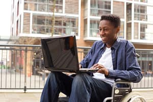 A young man sits in a wheelchair with a laptop computer in his lap. He is african american, smiling, and wearing jeans, a white shirt, with a denim shirt over it.