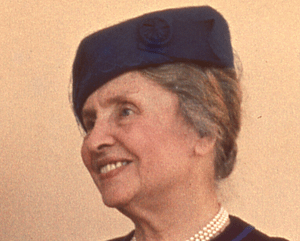 Helen Keller pictured, photo taken from the shoulders up. She is smiling, weating a dark blue pill box hat with her hair tied up on a tan background.