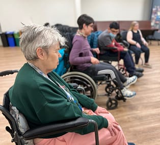 Consumers and an employee of CPWd enjoy the skills building and peer groups. Four people are lined up, the firs tthree in wheelchairs. The last woman in the row sits in a chair. They prepare for a seated yoga class.