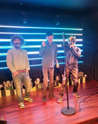 Three consumers dresses in costumes perform karaoke as part of the youth peer group karaoke night. They stand on a stage with a microphone and stand. Behind them is a wall with horizontal blue lights.