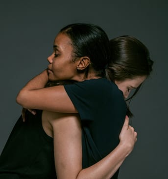 Two women hug. Both are wearing black t-shirts. One woman has black hair and dark skin. The other woman has brown hair and light skin.