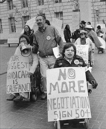 A black and white photo of demonstrators outside the San Fransisco HEW office building. In front of the group is Judy Heumann, a white woman who uses a wheelchair with short brown hair wearing glasses and a winter jacket with a “Sign 504” button pin. She is holding a sign that reads “NO MORE NEGOTIATION Sign 504.” To the left is Kitty Cone, a white woman who uses a wheelchair with short curly brown hair wearing glasses and a sweater. She is holding a sign that reads “OUR BIGGEST HANDICAP IS CALIFANO.” There are various other people around them holding signs and a mega phone.