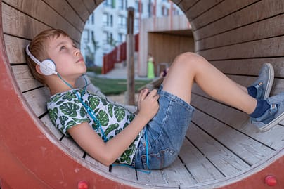 Smiling boy with smartphone and headphones listening to music outdoors, inside a circular wooden structure. He is wearing jean shorts and a patterned shirt. 