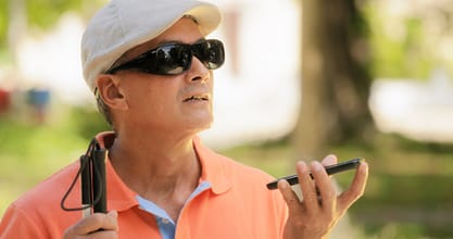 Image description: A visually impaired man is using Digital Assistant and Ease of Access functions on mobile phone, voice typing to smartphone. He is wearing sunglasses and a white hat, and holds a white cane in his opposite hand. 