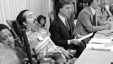 In this black and white photo, Ed Roberts sits in his wheelchair at a table. IN his mouth is a breathing tube. He has a strap across his chest. Several other people sit around him, such as men in suits and other people behind observing.