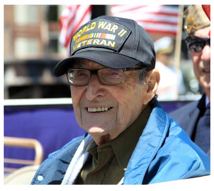 A senior man is smiling at the camera, pictured from the shoulders up. He wears a Worl War II veteran's baseball cap, and has a blue windbreaker on. Behind him waves an American flag.