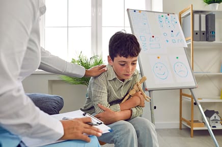 A child sits in a psychologist's office. He looks sad and is holding a wooden posable fugire doll. We see a white board behind him with different facial emotions drawn on it. The psychologists sits to the left of child. We only see thier arms and knees. One hand is placed on the child's shoulder. They have a clipboard in their lap. 