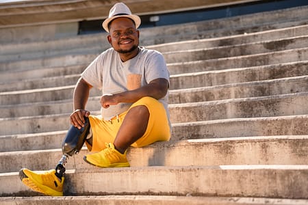 Image description: A black man wearing a white hat, shirt, and yellow shorts with prosthetic leg sits on some outdoor step, smiling at the camera. 