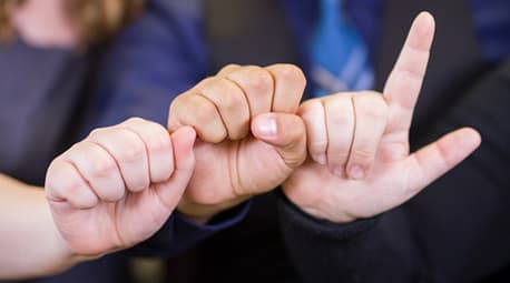 Image description: Tactile communication. Three hands are pictured up-close. They are touching side-by-side. The two hands on the left are in a fist, and the one on the right is in a fist with the index finger extended. 