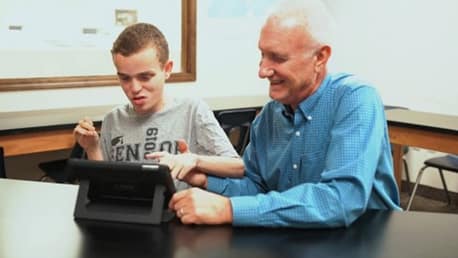 Learn4Life graduate John B. received personalized support to help him achieve a high school diploma. A young man sits next to an older gentleman who is guiding his hand to the screen of a tablet on a stand. (Photo: Business Wire)