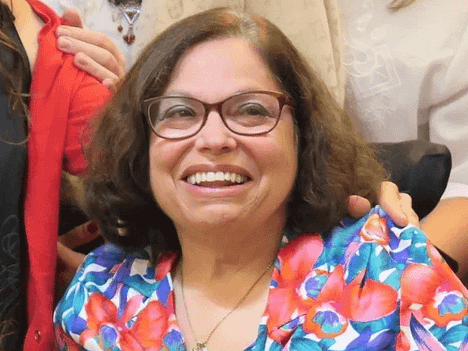 Activist Judy Huemann smiles in a headshot from the shoulders up, wearing a floral patterned shirt and brown-rimmed glasses.