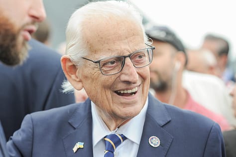 Congressman Bill Pascrell, Jr is seen here smiling, looking off camera. He has white hair, glasses, and is wearing a gray suit with a blue striped tie. 