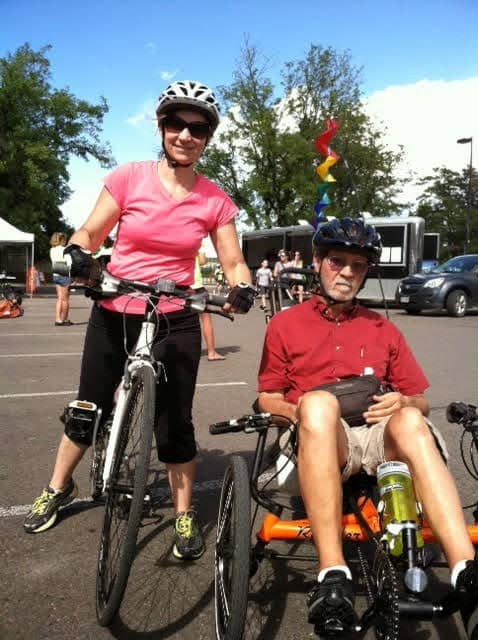Ed Milewski and his daughter Kristine are outside. Kristine is straddling a bike, wearing a helmet, sun glasses, a pink t-shirt and black capris. Ed is seated on an adapted orange bike, wearing tan shorts, a red shirt, sunglasses, and a helmet. 