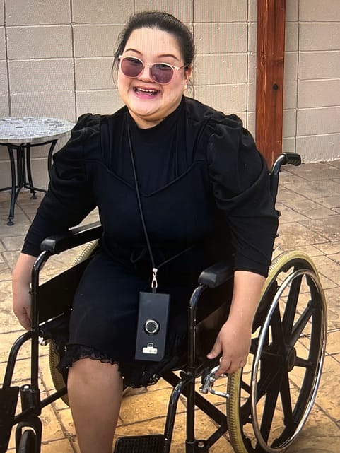 Image description: Mind Prawatsrichai sits in her wheelchair, smiling at the camera. She wears a black dress and sunglasses. Her black hair is pulled back. She sits in an outdoor courtyard.