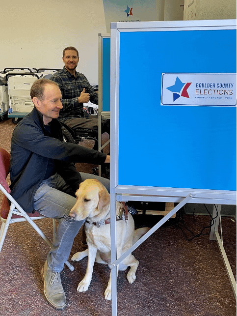 CPWD board member Michael Stone, who is blind, sits at a voting machine. His guide dog, a yellow lab, sits at his feet. In the background, Craig Towler, CPWD's Community Organizer, looks on smiling while giving a thumbs up. He sits in his wheelchair in front of another voting machine.