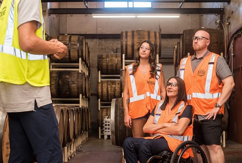 Three people wearing orange construction reflective vests sit and look at someone else to the left, who wears a yellow reflective vest. The woman in the front is in a wheelchair.