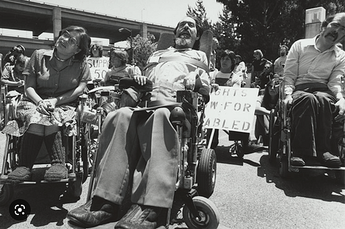 Ed Roberts and Judy Huemann. Image description: The photo is in black and white. Ed Roberts sits on the right in a wheelchair. He wears dark pants and a white shirt. Straps are across his torso in multiple places and his head is resting against a pad. To his left is Judy Huemann. She also sits in a wheelchair. She is wearing glasses with shoulder-length hair. She is wearing a button-down collared shirt and a skirt. She has on knee high knitted boots. Behind them are several other people, some holding signs that say "rights for disabled"