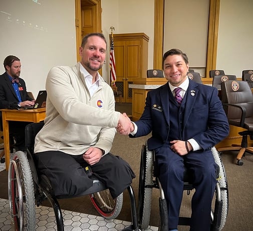 Craig Towler shakes the hand of representative David Ortiz, the sponsor of HB 23-1136. Image description: Both men sit in wheelchairs shaking hands and looking at the camera, smiling. Craig, seated on the left, wears a tan sweater over a white collared shirt and black pants. He has brown hair and a beard. His pants are folded underneath him at the knee where his legs end. Rep. David Ortiz is seated right and is wearing a blue suit with a striped purple and pink tie. Behind them is a man at a desk looking at a laptop. There is a cabinet and several brown chairs.