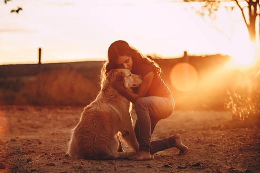 Image description: A woman crouches down and hugs her golden retriever emotional support animal. The sun sets and likes the scene in a golden color.