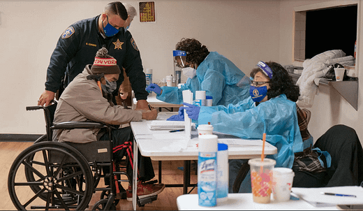 Preparing for a coronavirus vaccination in Essex County, N.J. A person in a wheelchair wearing a face mask and a ski hat sits in front of a table with two nurses in PPE. Credit...James Estrin/The New York Times
