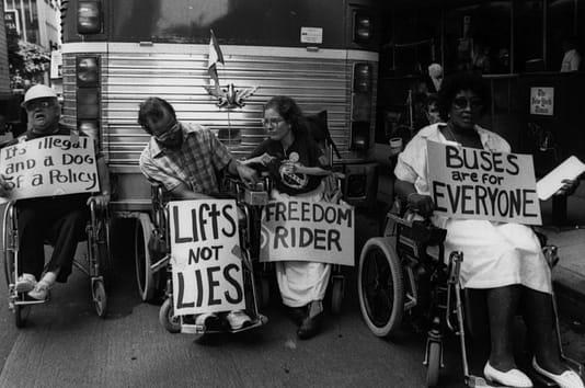 Disability rights activists stage a protest in Los Angeles, California, to bring attention to the inaccessibility of public transit and over-the-road coach companies in the 1970s. Image description: black and white image. Several people in wheelchairs sit in front of a public bus with protest signs. Photo Tom Olin photography.