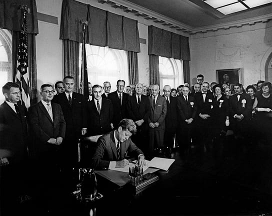 President John F. Kennedy signs the President's Panel into existence. Image description: The photo is in black and white. President Kennedy sits at a desk and is signing a document. He is wearing a dark suit and tie. Standing behind him are dozens of men and women in suits and dresses watching. Behind the crowd of men and women are large windows with paneled drapes.